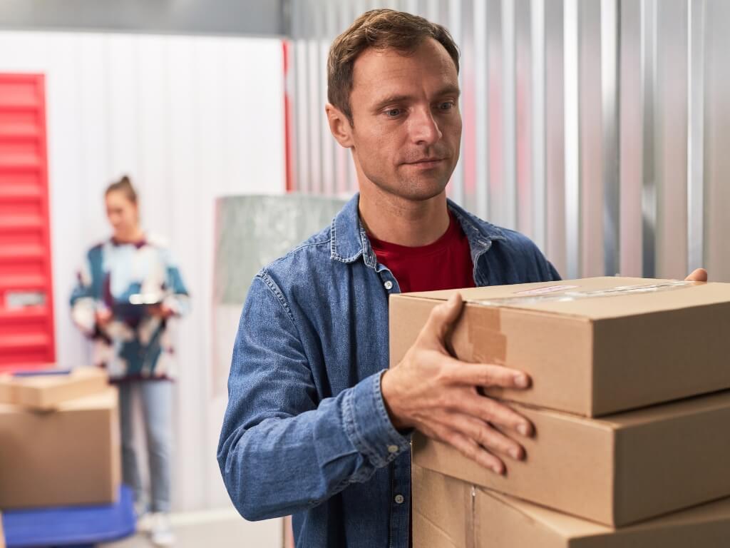 A man carries a stack of boxes in a self-storage room, organizing his belongings efficiently.